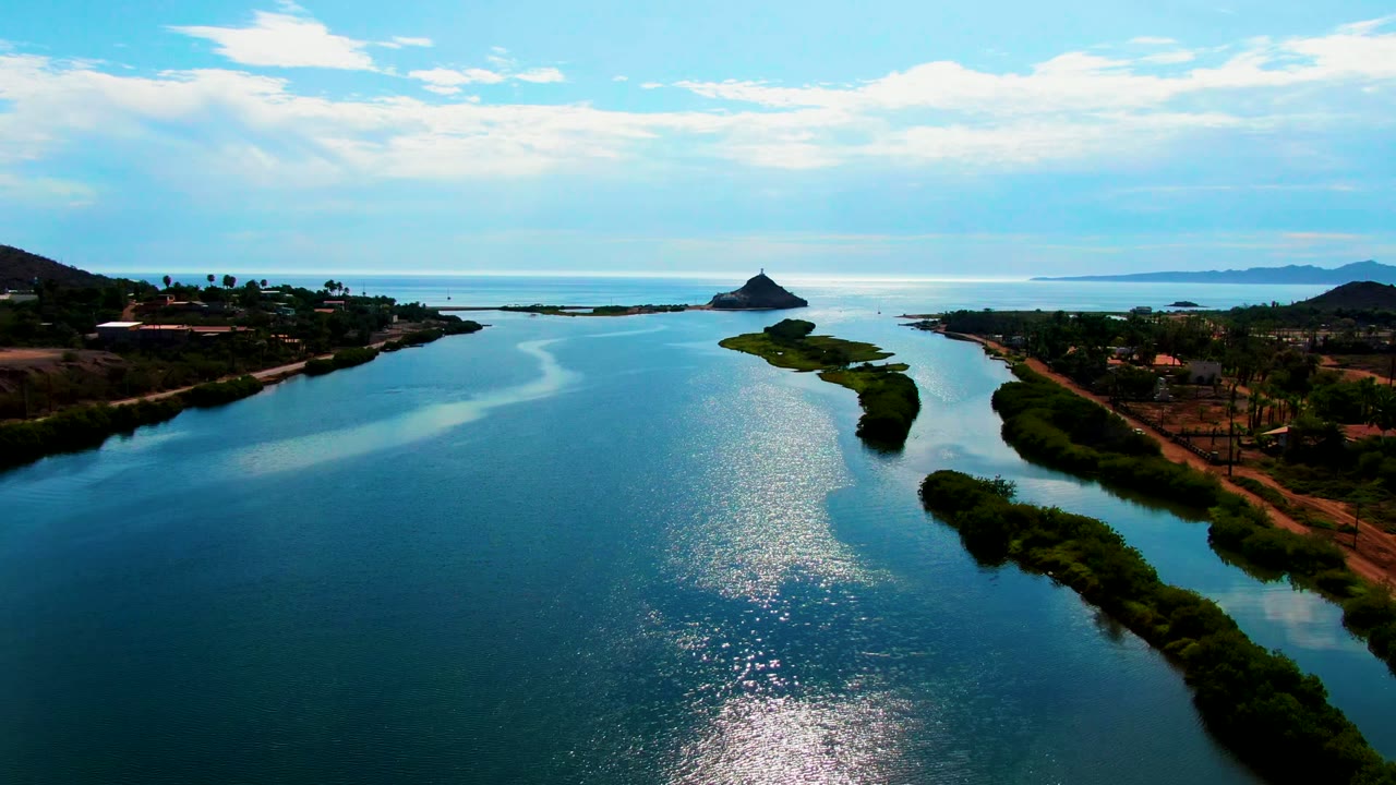 A Mulege aerial frame centers the reflective river surface with island vegetation to the right, low coastal banks and roads along the edges, a pointed hill near the horizon, cloud cover, and no visible people.