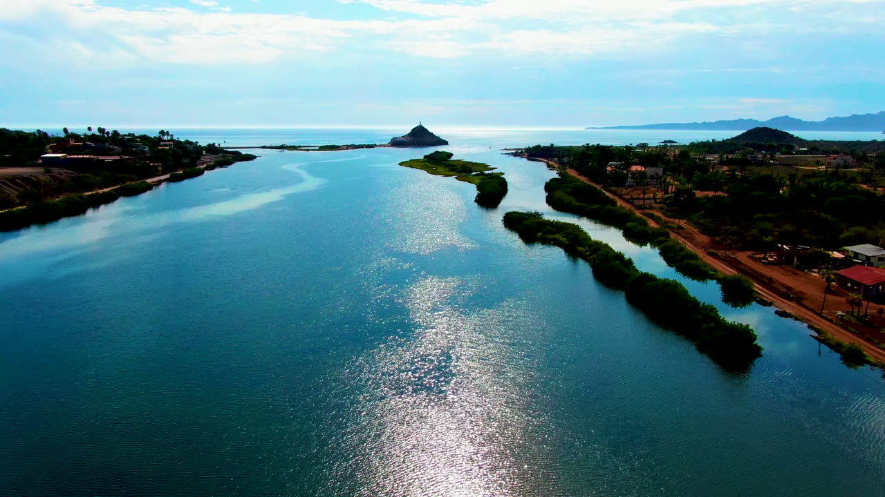 Opening aerial frame over a Mulege, Baja California Sur waterway, with low land on both sides, distant hills, bright sun reflection on the blue-green surface, and no visible people.