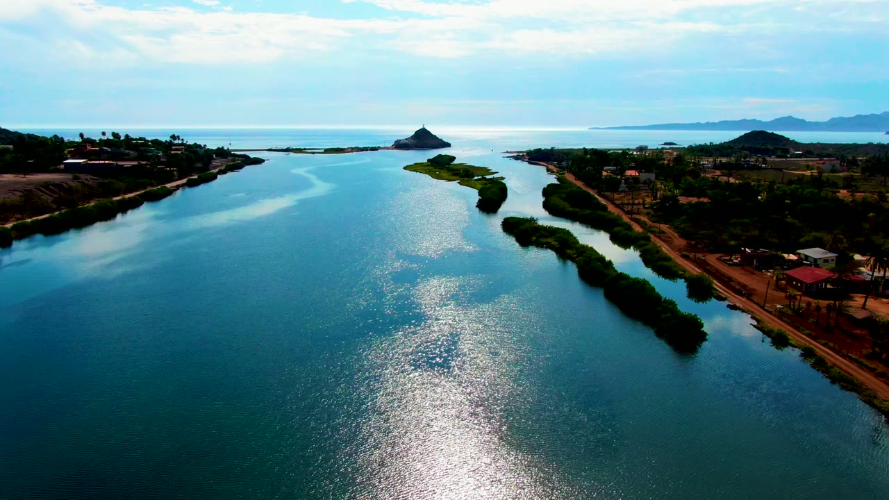 Mid-clip aerial view over the Mulege waterway, with a bright shimmer crossing the foreground water, low banks to either side, scattered buildings, distant hills, and no visible people.