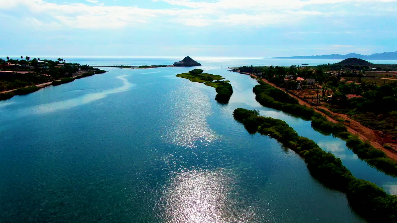 Aerial river view with shimmering water, green shoreline, small island, and low buildings near the bank.