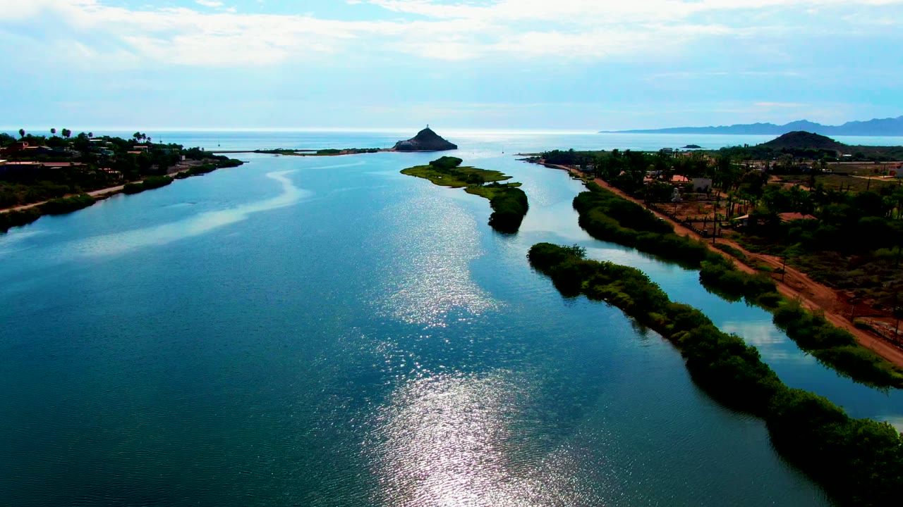 Third frame of the Mulege river view with the small island near the channel mouth, darker green banks at right, open blue water in front, distant mountains, and no visible people.