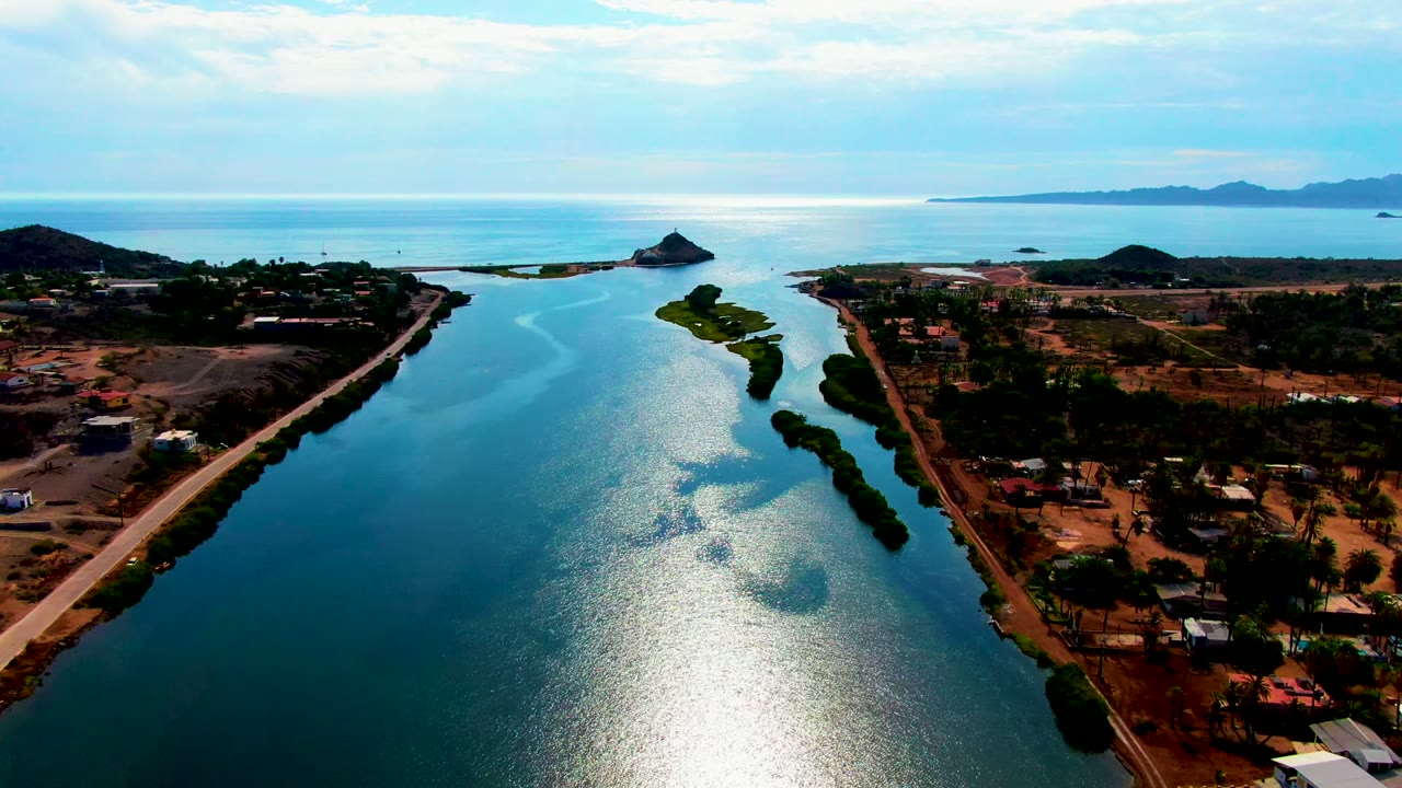 High aerial frame over Mulege, Baja California Sur showing a blue canal, roadside settlement, small green island, distant ocean and hills, and no visible people.