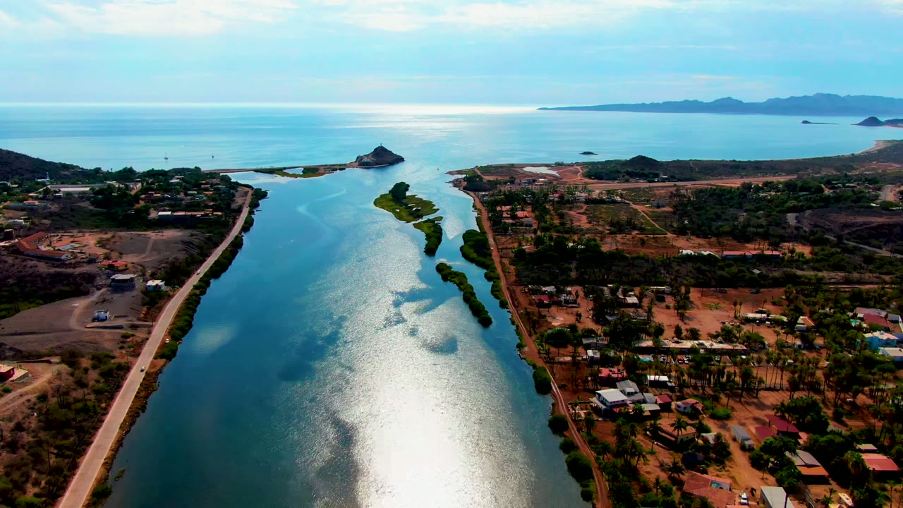 Opening aerial frame over Mulege, Baja California Sur, with a blue canal leading to the ocean, a road along one side, scattered buildings, trees, and no visible people.
