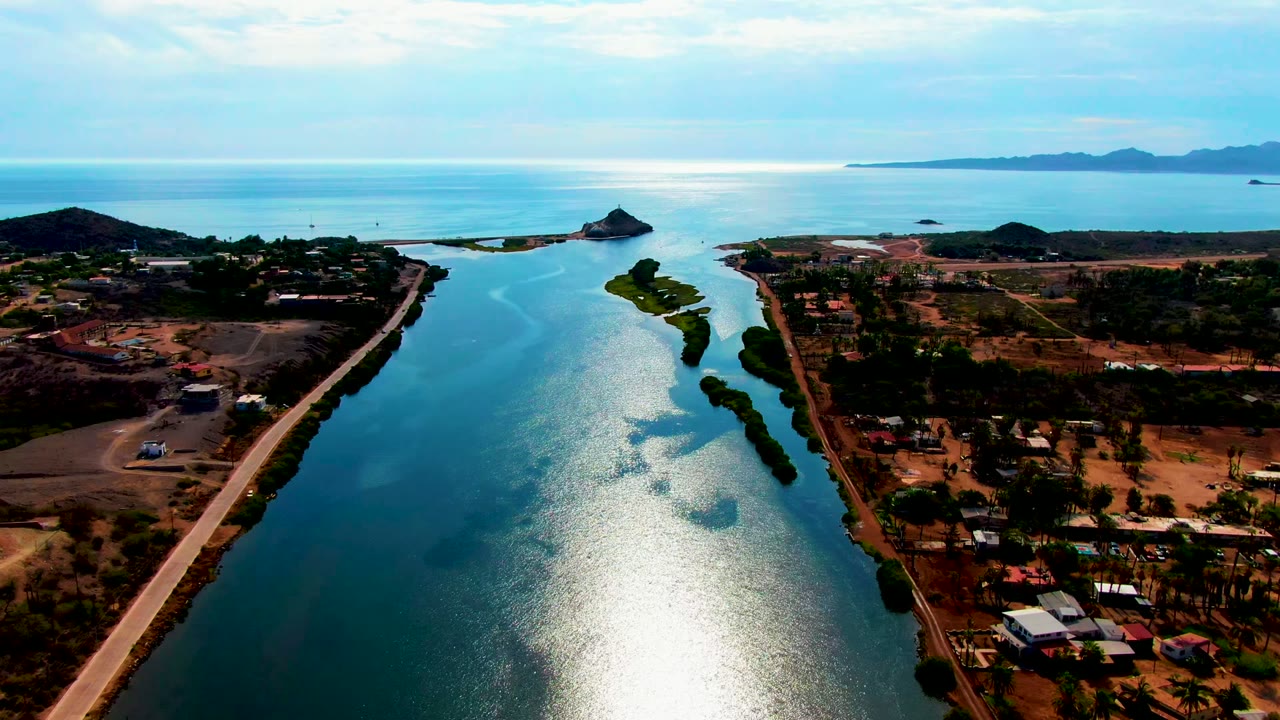 Aerial frame over Mulege, Baja California Sur showing a narrow reflective waterway, road, dry coastal plots, scattered buildings, ocean, distant hills, and no visible people.