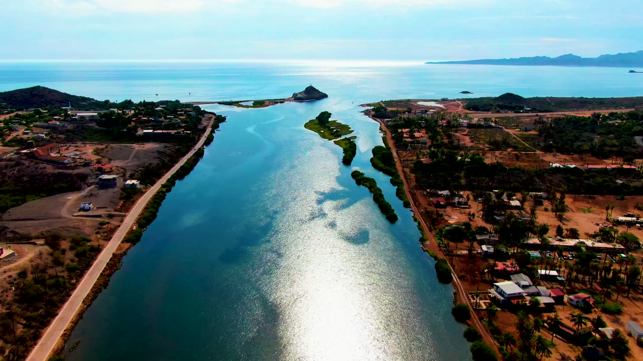 High aerial view of a sunlit channel with road traffic and low hills beyond.