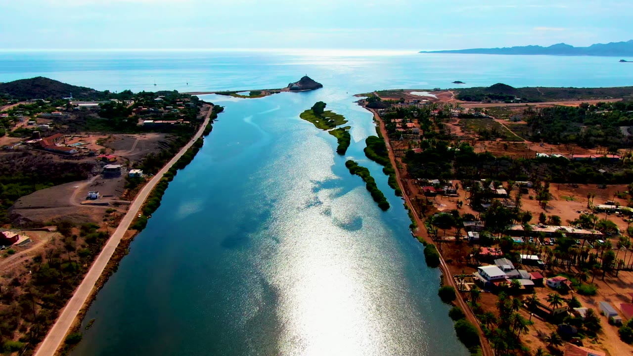 Mid-clip aerial over Mulege shows the waterway centered below the drone, a pale road tracking the bank, dry lots with buildings, offshore landforms, distant hills, and no visible people.