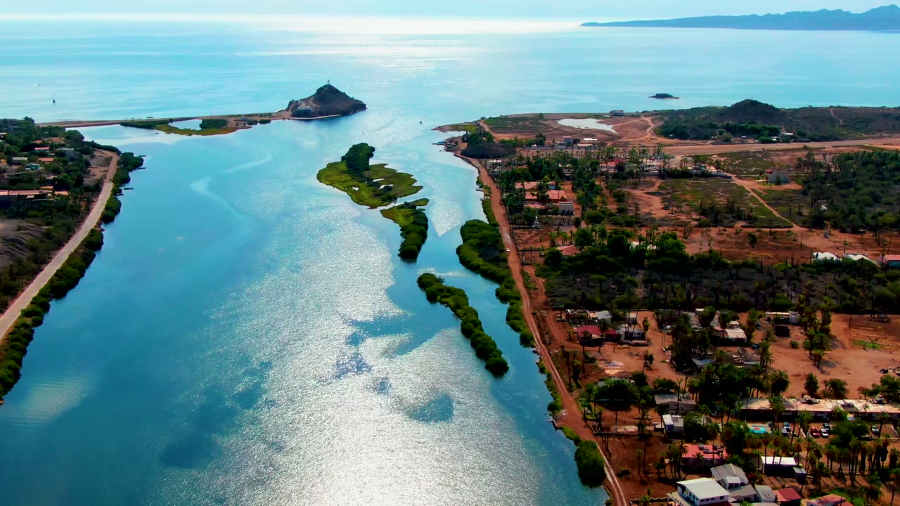 Opening aerial frame over Mulege, Baja California Sur, with bright river water on the left, a green island near center, settlement and road on the right, and no visible people.