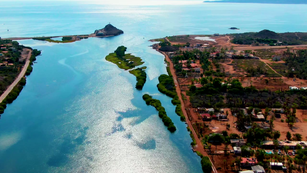 Aerial river scene with reflective water, small island, shoreline road, buildings, and distant hills.