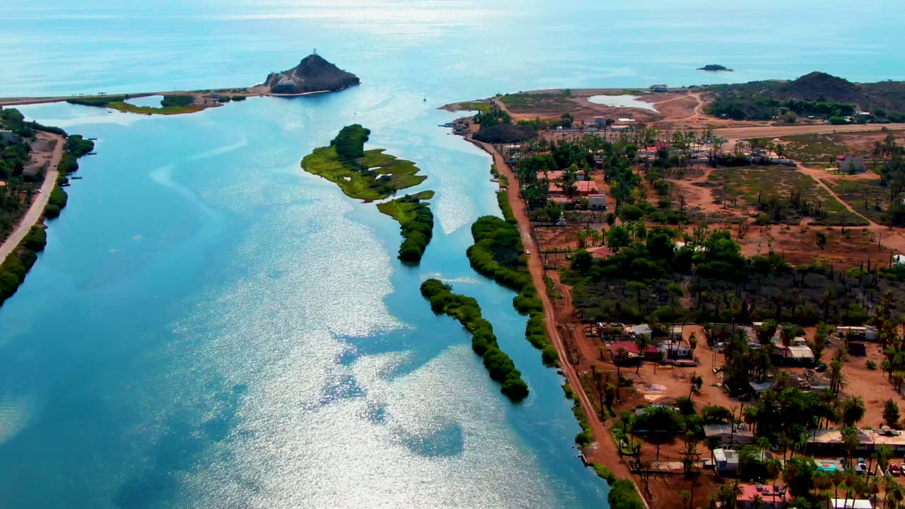 Mid-early aerial frame over Mulege, Baja California Sur, with more of the right-bank settlement visible beside the river, the green island below the horizon, and no visible people.