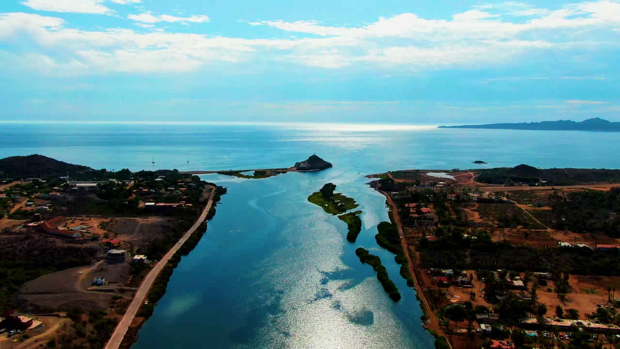 Aerial frame over Mulege, Baja California Sur, showing a blue-green canal between dry land and sparse vegetation, with open ocean, distant hills, clouds, and no visible people.