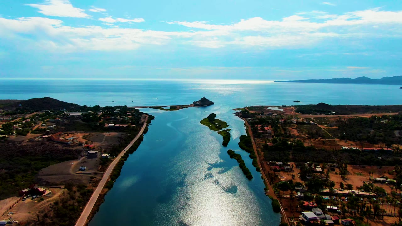 A high aerial frame over Mulege, Baja California Sur, showing blue water cutting through brown coastal land, a road along the right side, low vegetation, distant ocean, and no visible people.