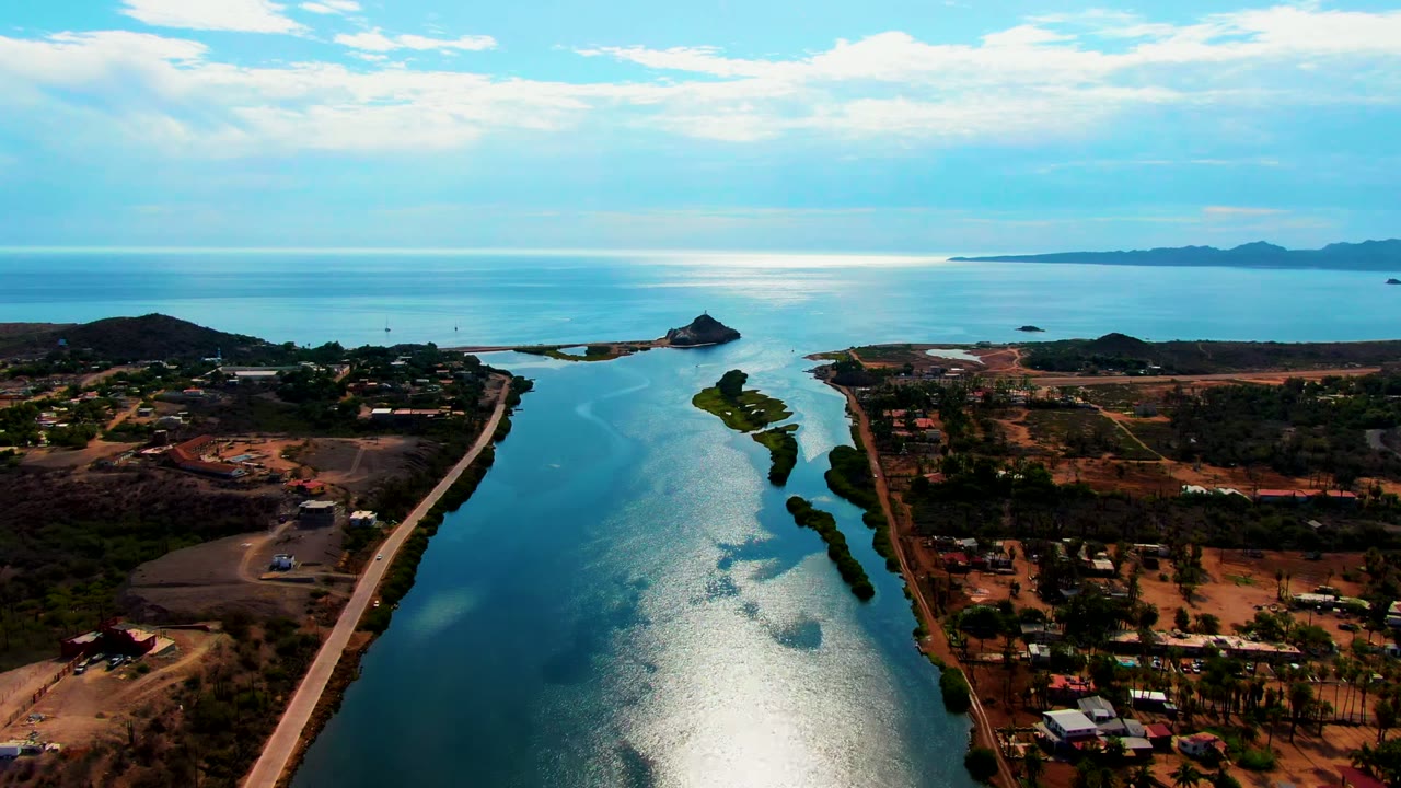Aerial frame over Mulege, Baja California Sur showing a blue waterway leading toward the ocean horizon, roads and scattered buildings on dry land, clouded sky, and no visible people.