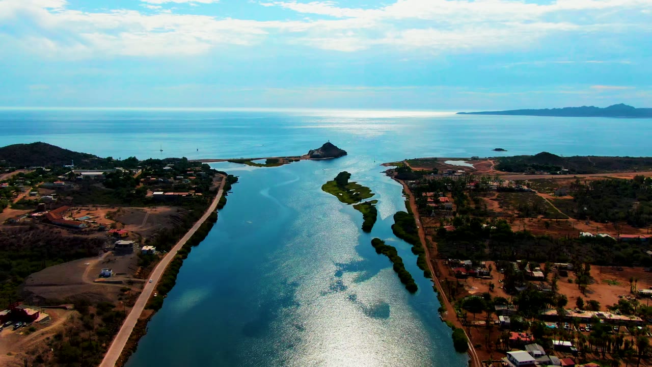 Aerial view over Mulege, Baja California Sur with the canal-like waterway running toward bright ocean water, dry shoreline lots, a roadside strip, small structures, low clouds, and no visible people.