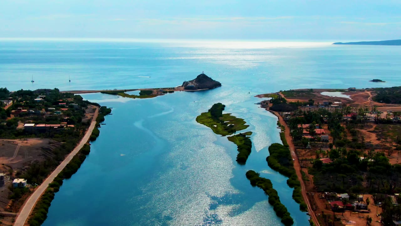 Aerial frame over Mulege, Baja California Sur, showing a narrow turquoise channel, shoreline roads, scattered buildings, offshore rocks, ocean horizon, and no visible people.