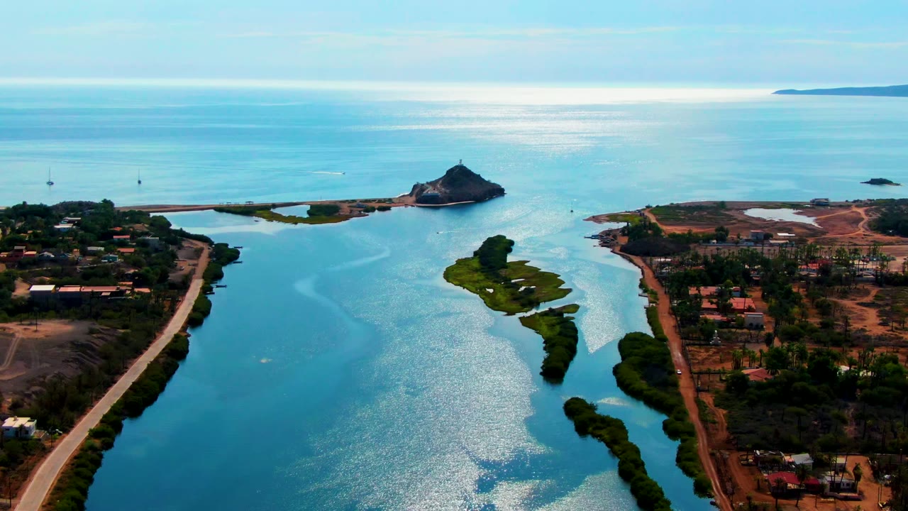 Elevated Mulege coastal frame showing the channel running toward open water, roads and low buildings on dry land, small boats or masts in the distance, and no visible people.