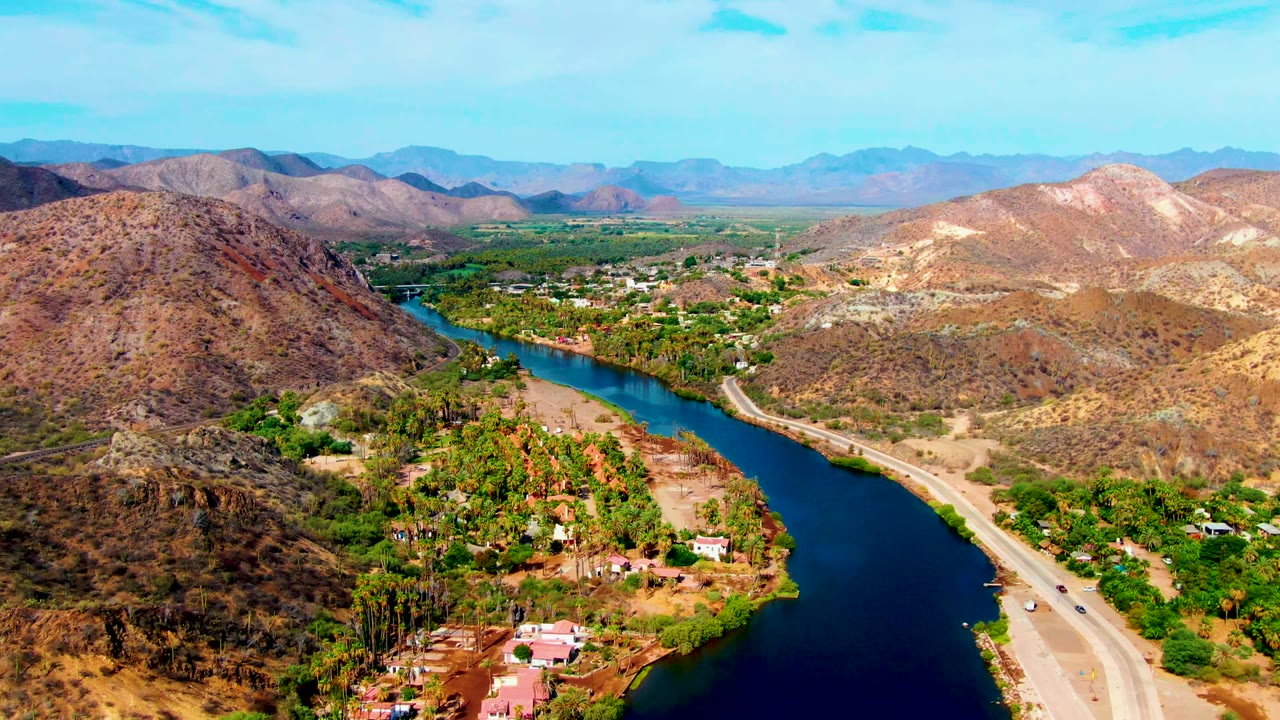 Aerial view over Mulege, Baja California Sur, with a dark blue river curving beside a pale road, green banks, scattered homes, dry hills, and no visible people.