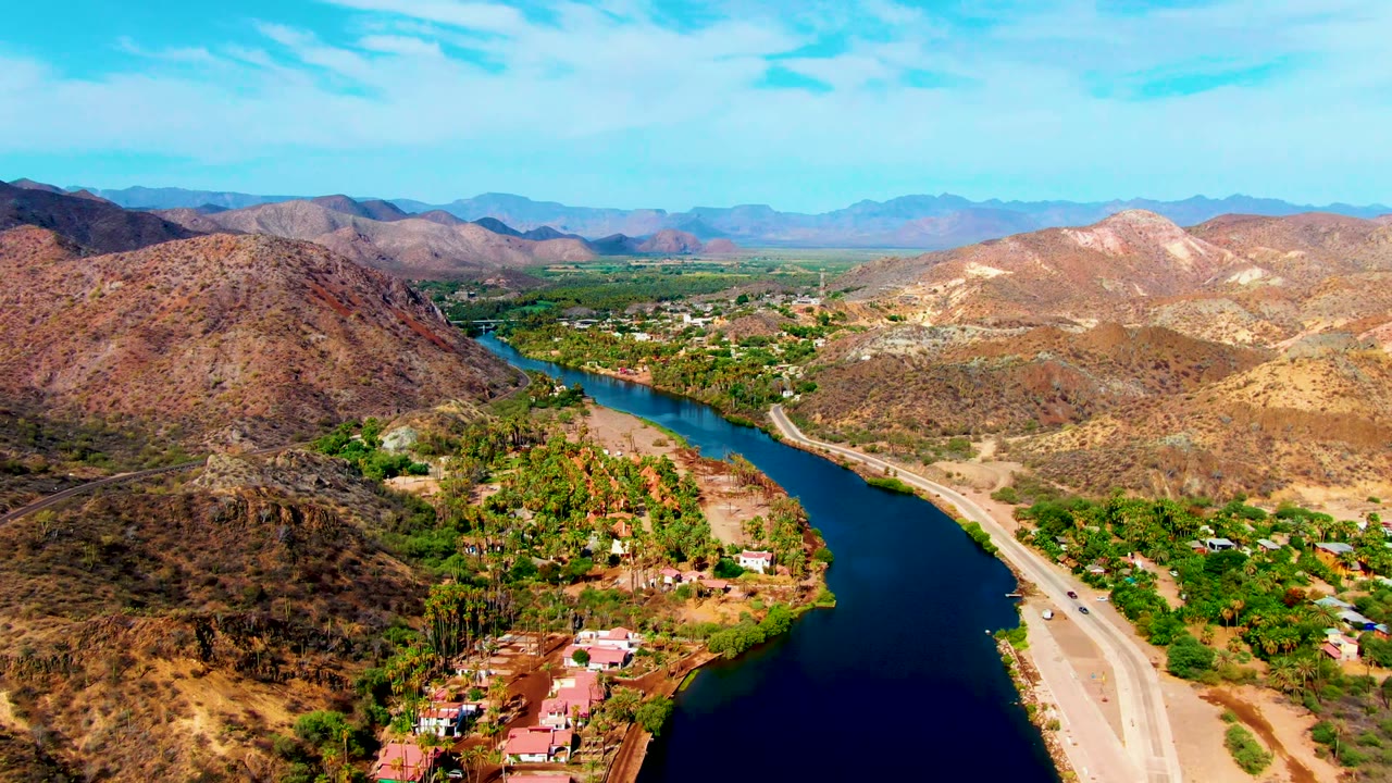 Wide aerial view in Mulege, Baja California Sur, with the river cutting between dry hills, a road carrying small vehicles, homes and palms near the water, and no visible people.