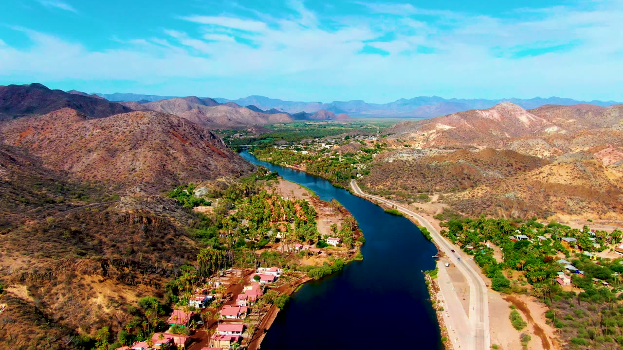 Opening aerial frame over Mulege, Baja California Sur, with a dark river curving past red-roof buildings, a pale road, dry hills, and no visible people.