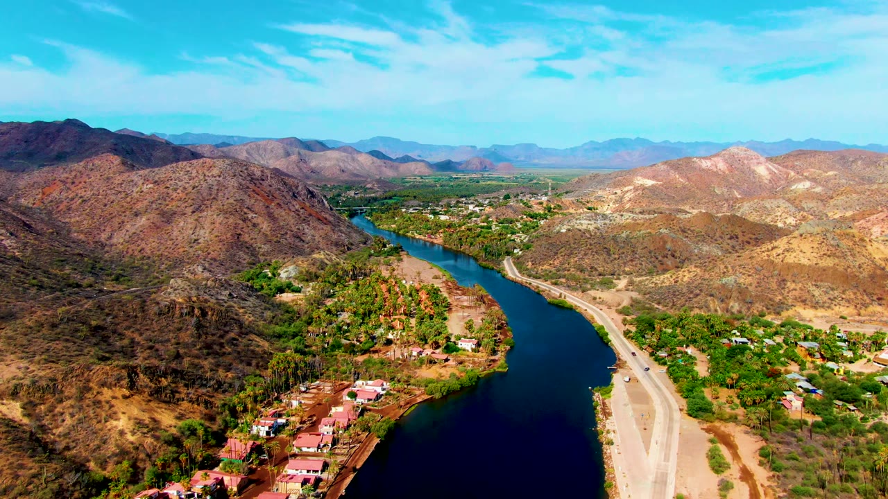 Wide drone frame over Mulege, Baja California Sur, with a dark blue river, red-roof riverside buildings in the lower frame, a light road on the bank, and no visible people.