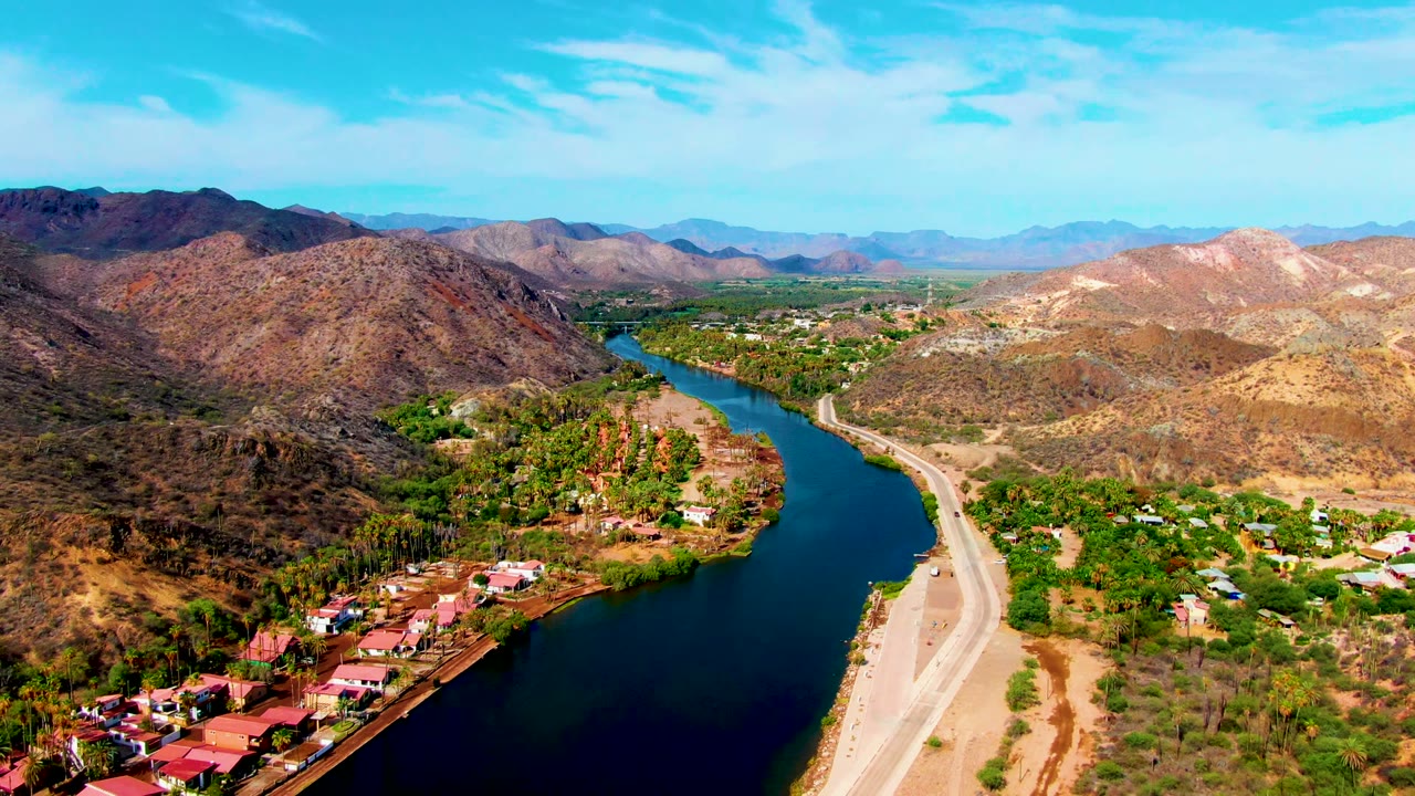 Aerial frame over Mulege, Baja California Sur, showing a blue-green river beside a pale riverbank road, red-roofed buildings, dry hills, and no visible people.