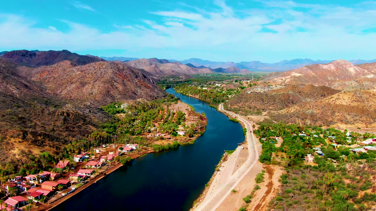 Mulege aerial frame showing a dark blue river bend, red-roofed riverside buildings, green trees, a road on the opposite bank, dry slopes, and no visible people.