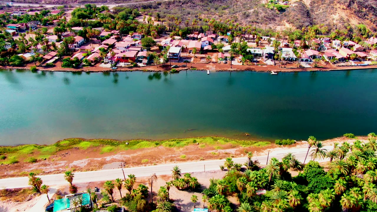 Aerial frame from Mulege, Baja California Sur, Mexico showing turquoise water across the center, dry exposed shore, a roadside strip, palms, homes, and hills, with no visible people.