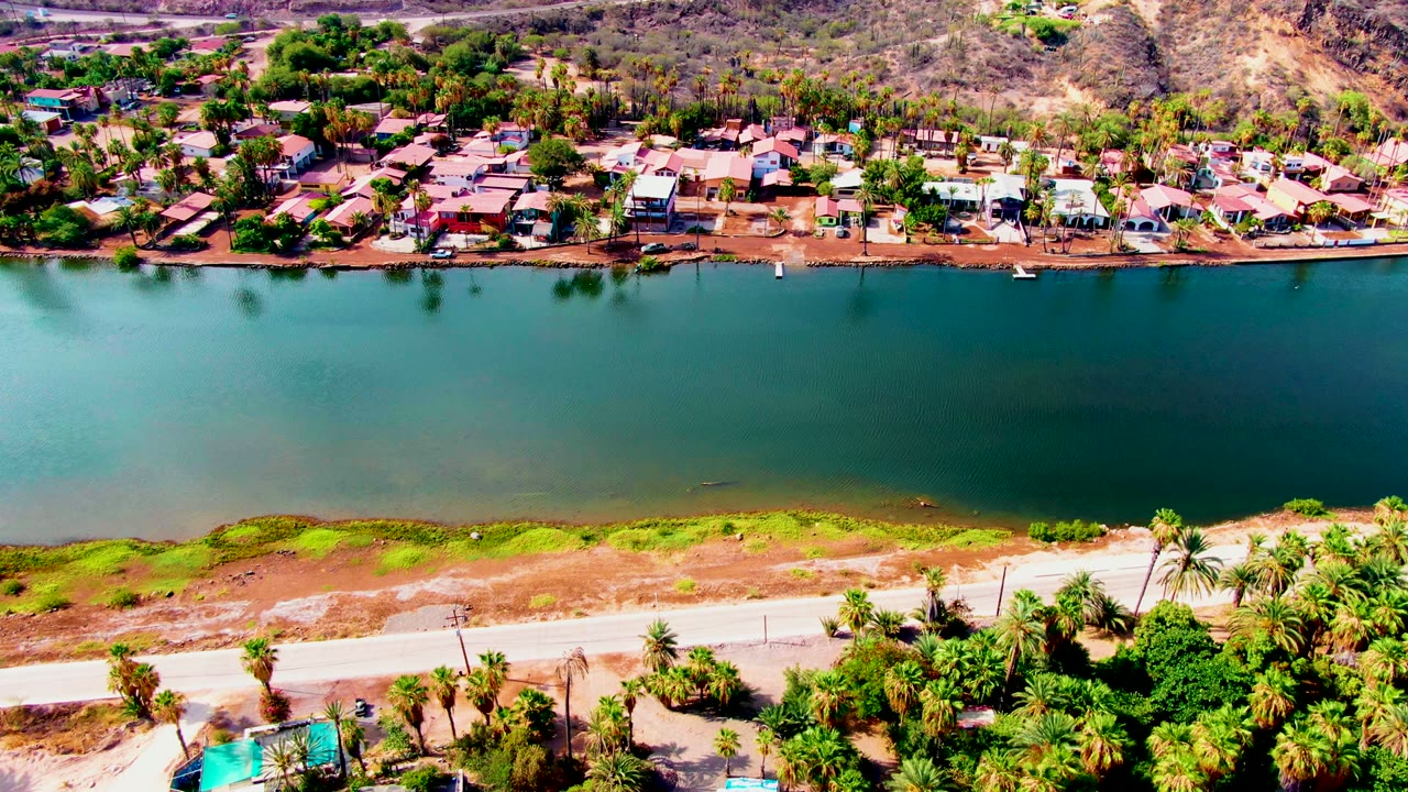 Drone view over water toward a palm-lined residential shore and hills.