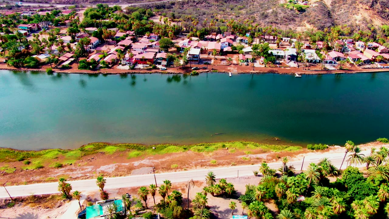 At 0:00, an aerial view over Mulege, Baja California Sur shows green reservoir water between a palm-lined road and a residential shoreline, with no visible people.