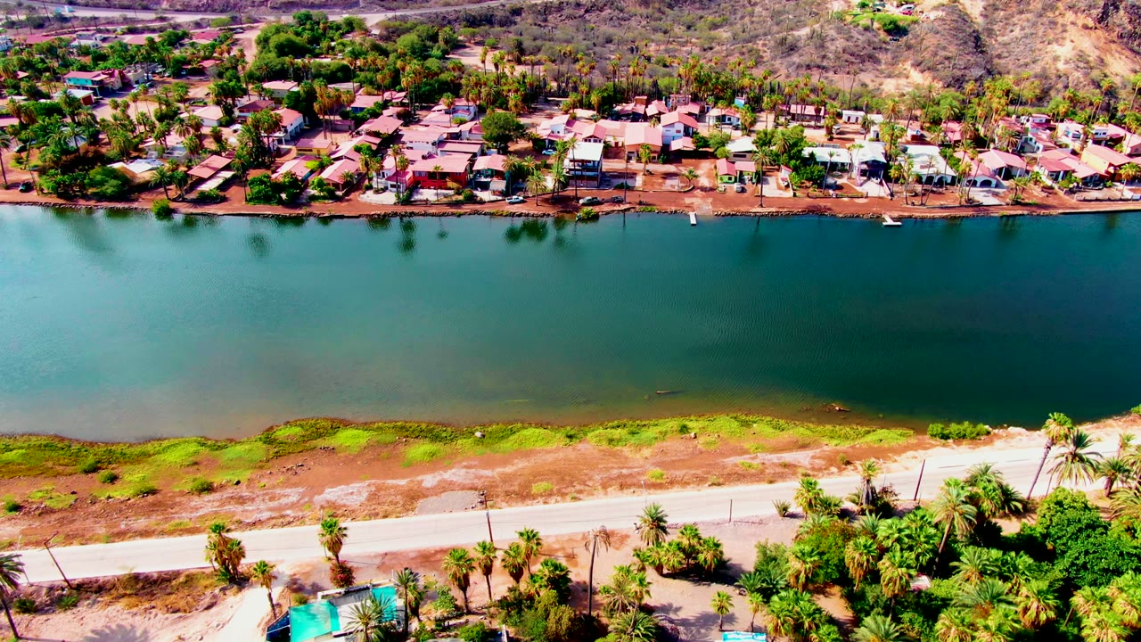 At 0:02, bright overhead light reveals the Mulege reservoir, a pale road with palms in the foreground, and a dense line of homes beyond the water, with no visible people.