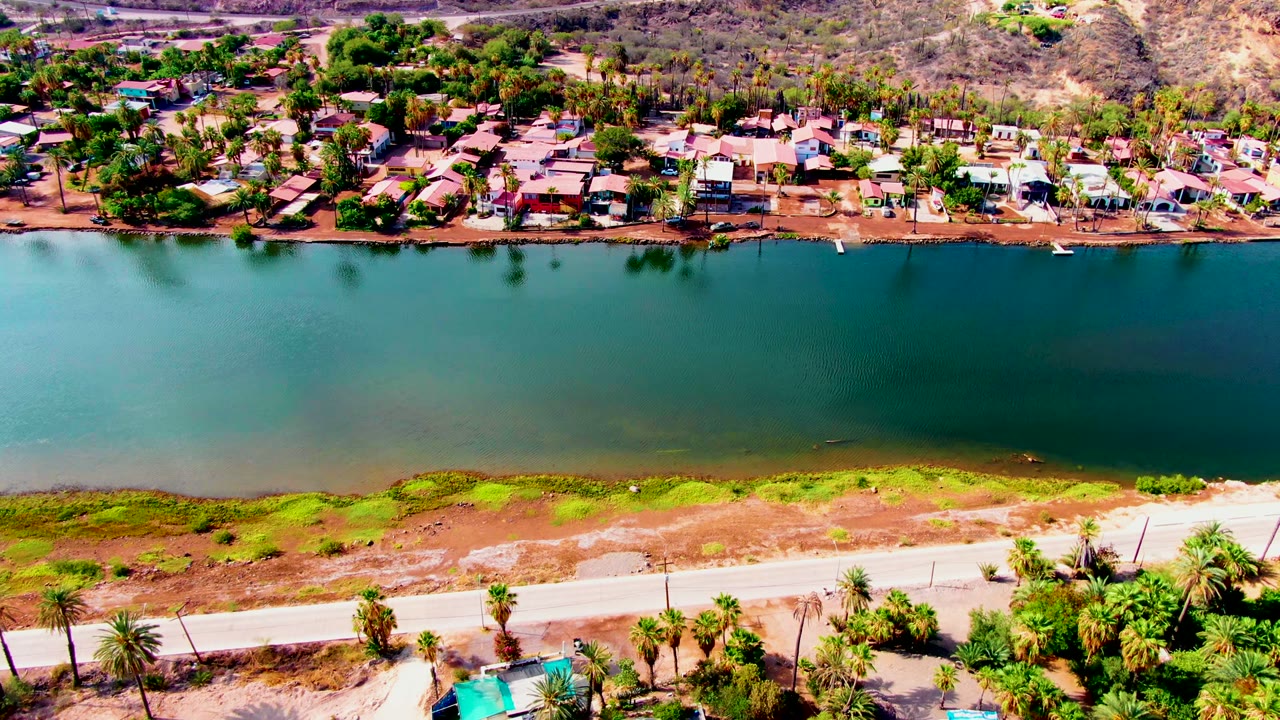 Opening aerial frame over Mulege, Baja California Sur with teal water centered between foreground road and background shoreline homes, with no visible people.