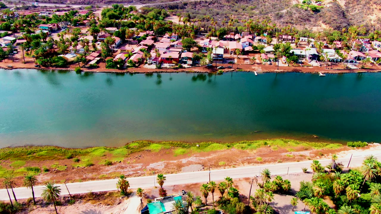 Wide aerial view of calm water with homes, trees, road, and palms.