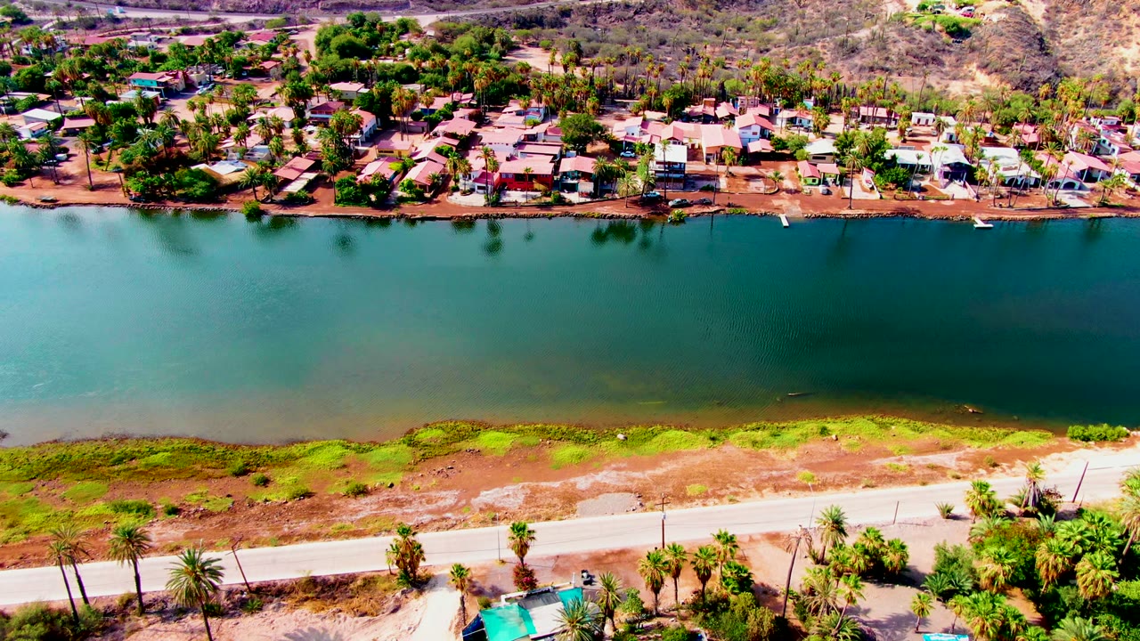 Third sampled frame over Mulege, Baja California Sur with teal water filling the middle, homes and trees along the upper bank, roadside palms below, and no visible people.