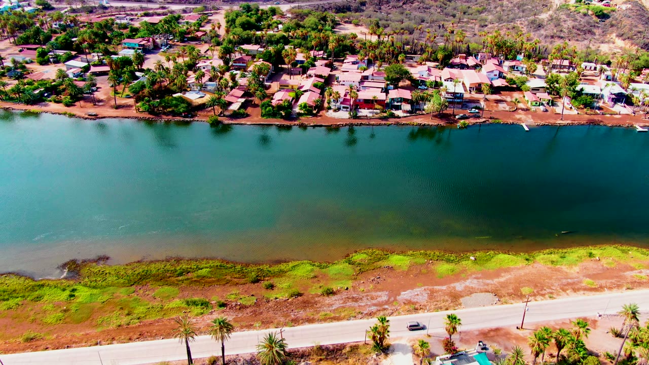 Aerial frame from Mulege, Baja California Sur showing a teal canal between a pale road and a residential shoreline, with a small car on the near road and no visible people.