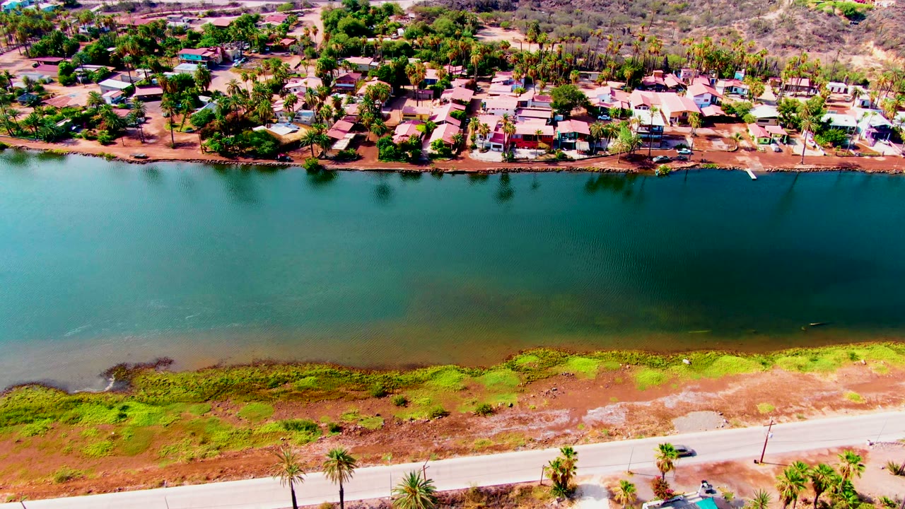 Aerial canal scene with a car farther along the road and homes across the water.