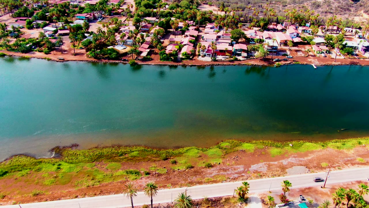 Mulege aerial frame showing the car near the right side of the road, a broad teal canal, green algae along the near shore, and dense homes and trees on the opposite bank with no visible people.