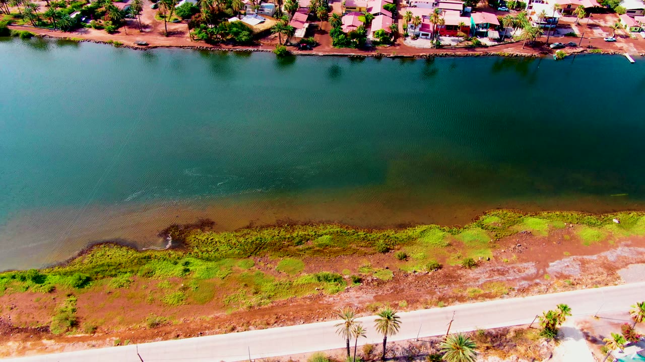 Aerial canal water beside a marshy bank and road with homes beyond.