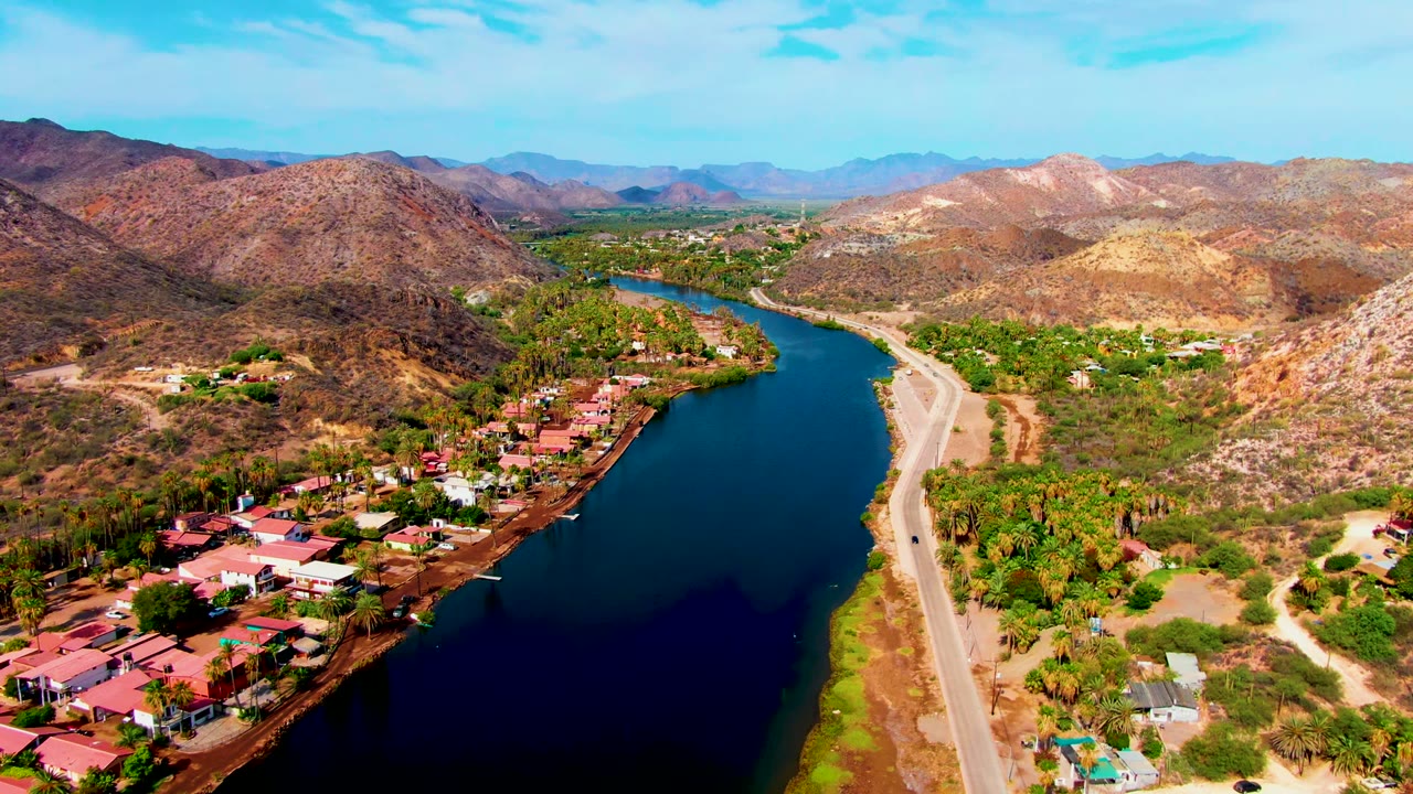 Aerial view in Mulege, Baja California Sur, with a dark blue river at left, a parallel road at center-right, red-roofed homes, dry hills, and no visible people.