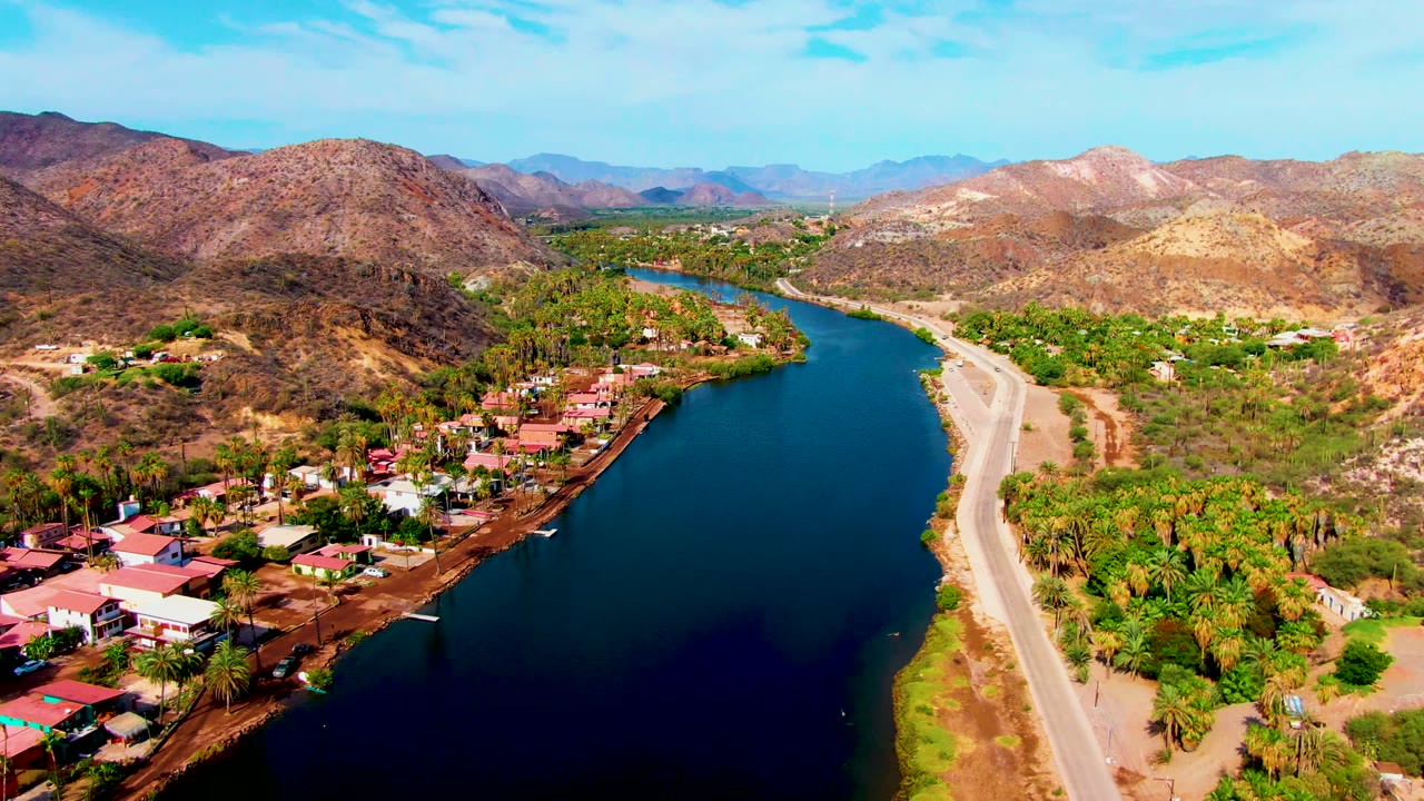 Aerial view in Mulege, Baja California Sur showing a blue river curving beside a pale road, red-roofed homes, trees, and dry mountains, with no visible people.