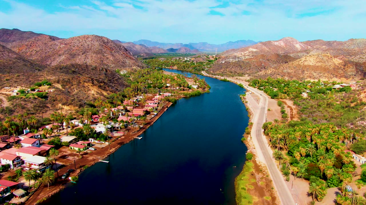 Wide aerial view of a curved river, roadside homes, green trees, and mountains.