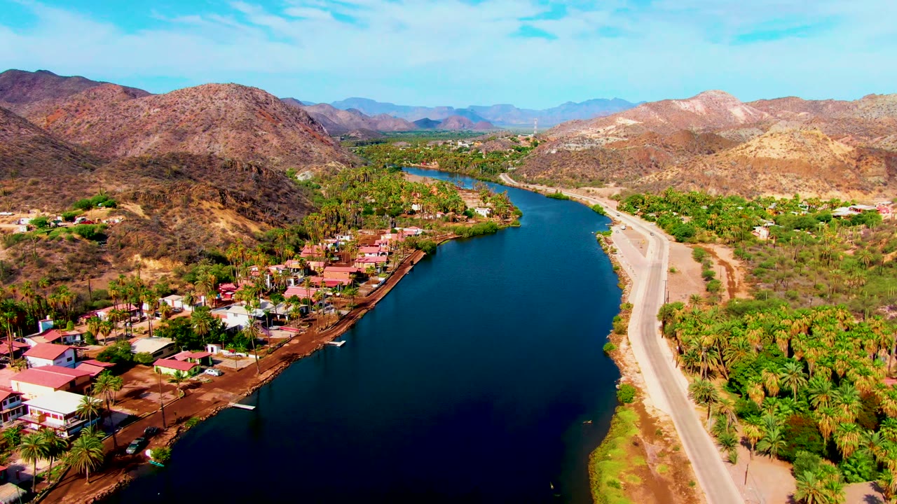Aerial view over Mulege, Baja California Sur showing a river bend, pale roadside, red roofs, palms, dry hillsides, distant mountains, and no visible people.