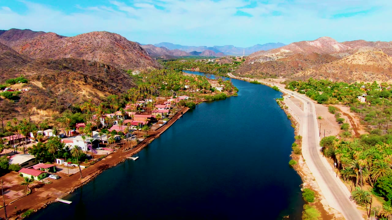 Aerial frame over Mulege, Baja California Sur showing blue river water, a riverside road, red-roofed homes, palms, dry hills, distant mountains, clear sky, and no visible people.