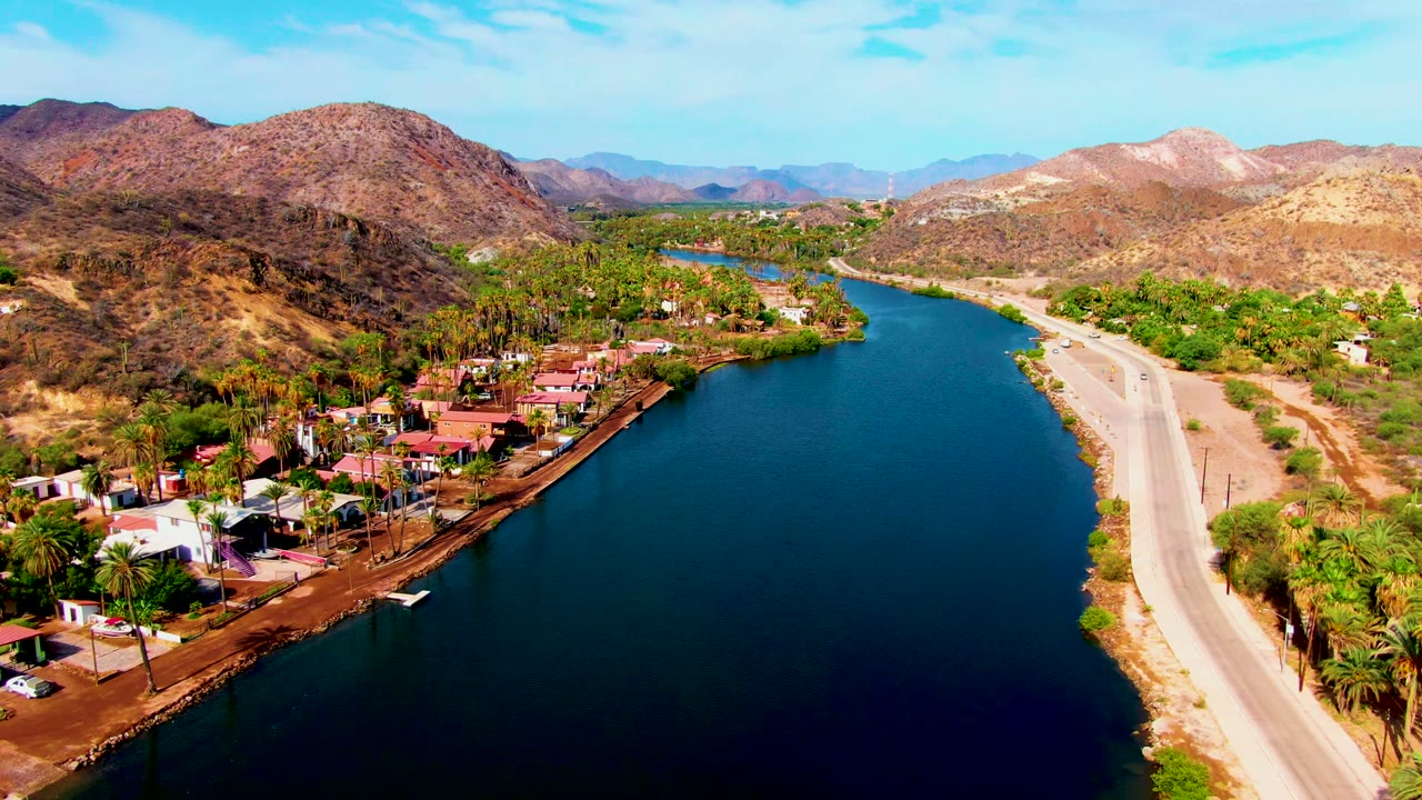The Mulege aerial view holds on blue water in the foreground, homes and palms along the left bank, a road on the right bank, layered dry hills, and no visible people.