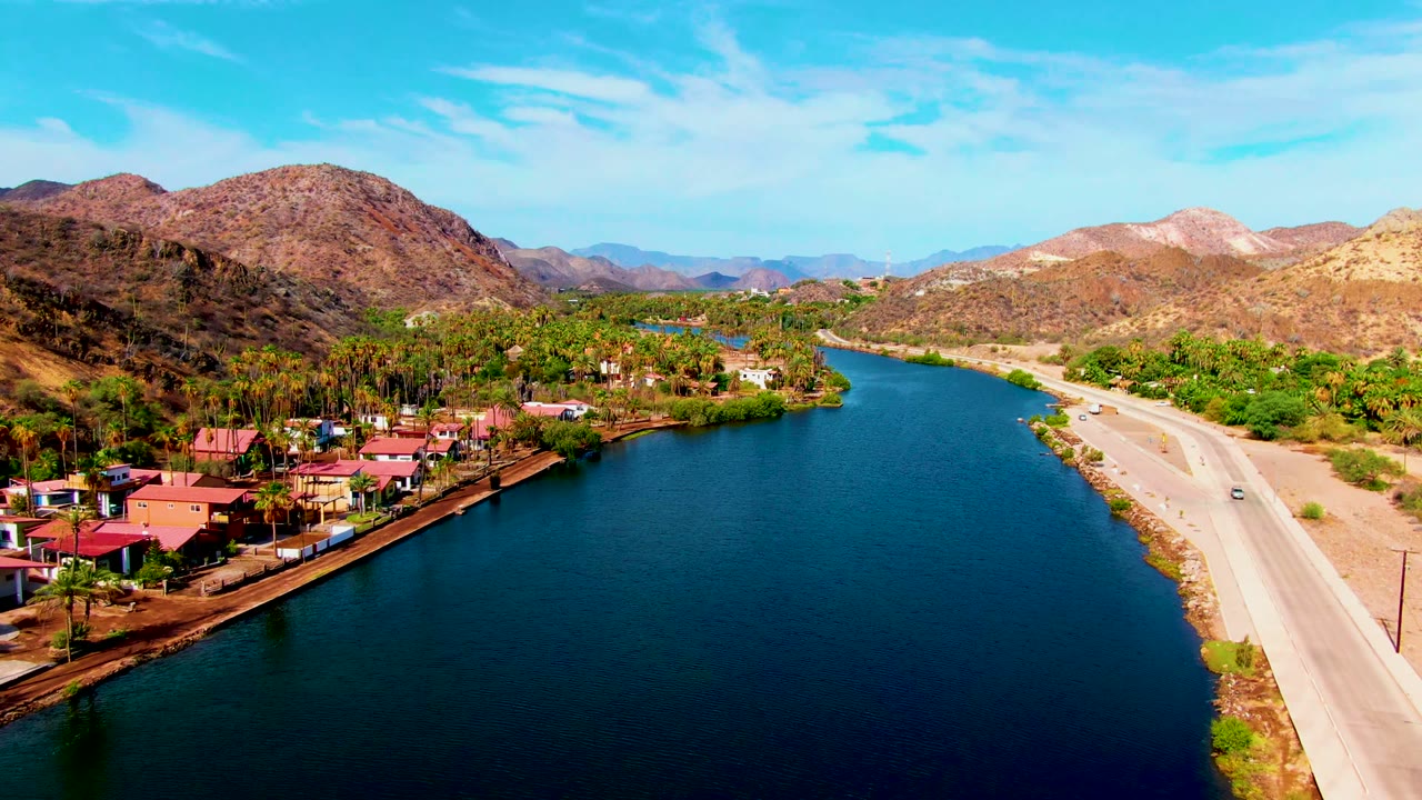 At the start of the Mulege, Baja California Sur clip, a blue river fills the center with a paved road on the right, red-roofed buildings on the left, dry hills beyond, and no visible people.