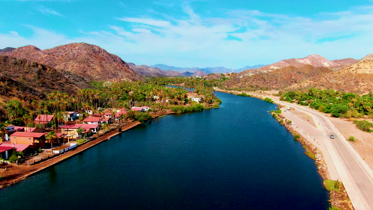 Wide aerial frame of river water, roadside buildings, and arid mountains.