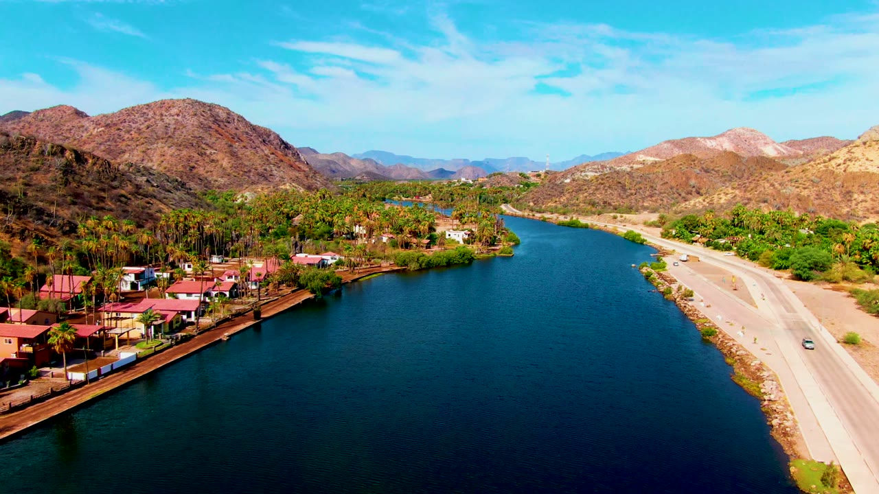 At 0:02, the aerial view looks down the Mulege river corridor with the road on the right, homes and palms along the left shoreline, a small moving car, tan hills, and no visible people.