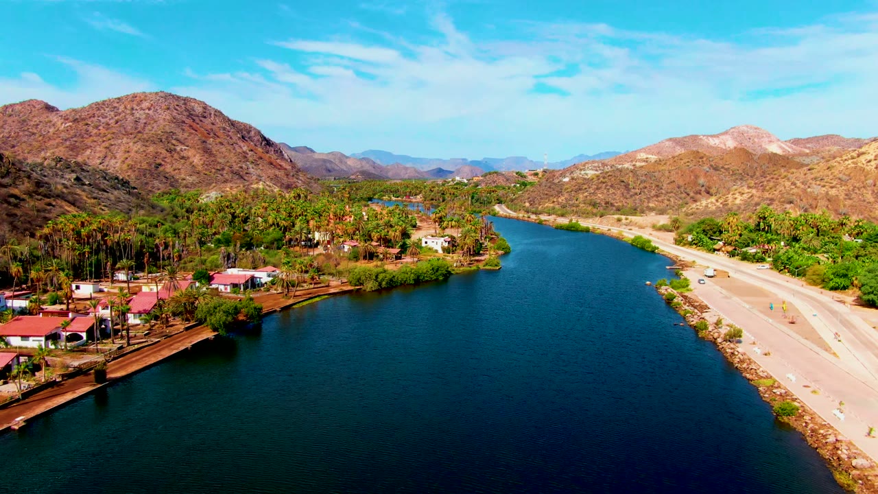 Opening aerial frame over Mulege, Baja California Sur, with dark blue river water in front, a light road along the right bank, palm trees and homes on the left, brown mountains behind, and no visible people.
