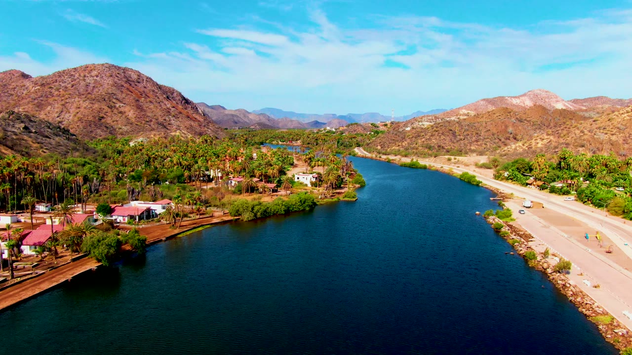 Wide aerial frame over Mulege showing blue river water across the foreground, the road and vehicles along the right edge, a green palm area with houses to the left, desert mountains beyond, and no visible people.