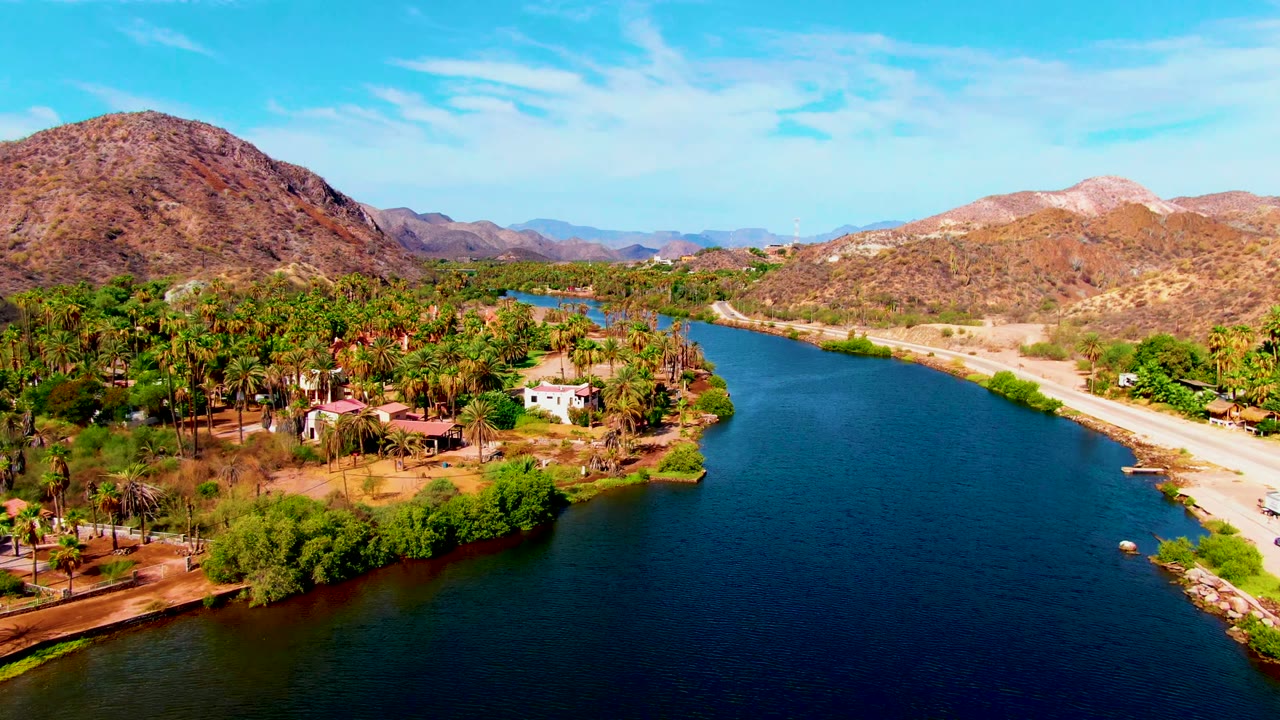 Opening aerial frame over Mulege, Baja California Sur, with a blue river curving past palm groves, small buildings, a pale roadside, desert mountains, and no visible people.