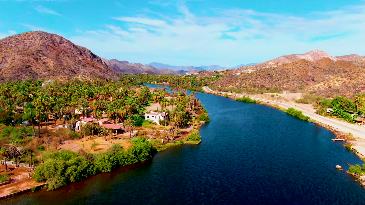Aerial river landscape with palms and buildings on one bank and a road below desert hills.