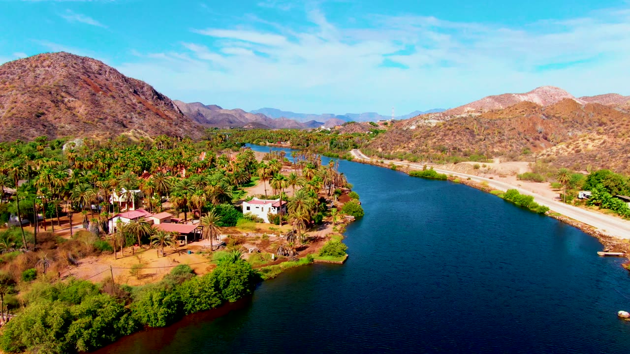 At 0:02, the Mulege river runs deep blue through the frame, bordered by palms and buildings on the left bank, a road on the right bank, desert mountains, and no visible people.